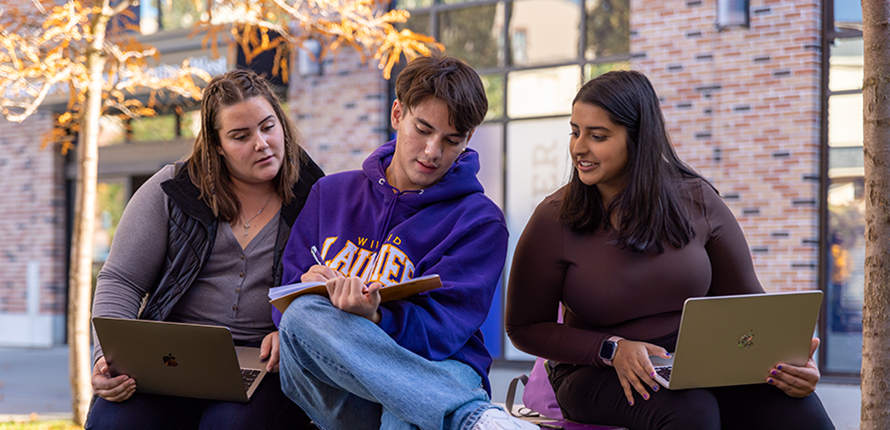 Three students sitting outside the Research and Academic Centre studying with laptops and notebooks