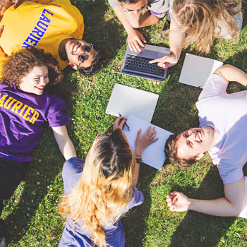 A student smiling at the camera, outdoors during the summer, wearing a Laurier shirt.